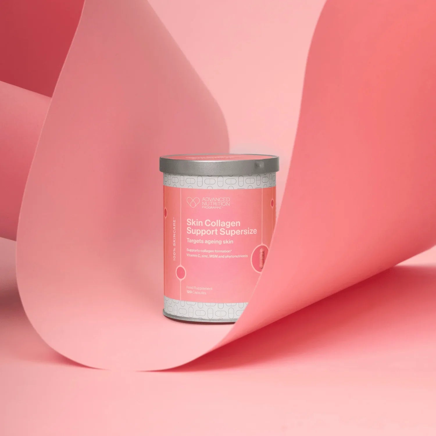 A cylindrical supplement tin labeled 'Skin Collagen Support Supercharge' by the brand 'Vitality', set against a pink studio background with a large pink paper scroll behind it.